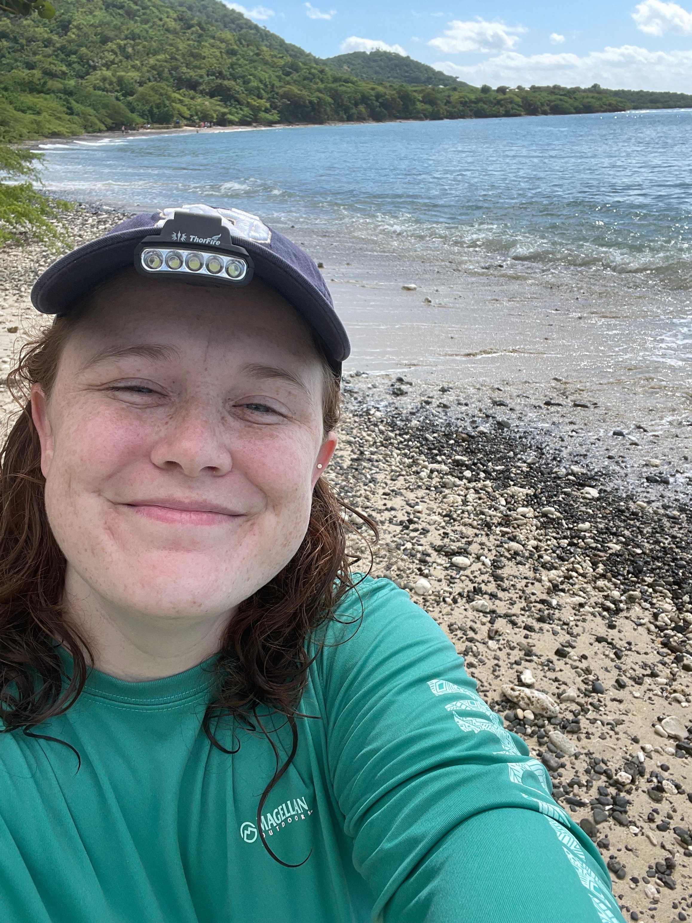 a selfie of Haley Barlow on a beach. Haley is wearing a navy blue baseball cap and a teal blue long sleeve shirt. she is smiling with her eyes closed. the ocean is to the right, and the trees on the beach are to the left of the image. 