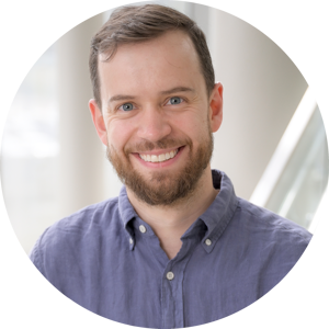 A headshot of Daniel Bondeson, wearing a smile, a beard, and a light blue, button-down shirt. The photo is set indoors with a white modern staircase railing in the background.