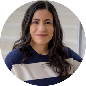 A headshot of Jennifer Vega with long dark hair in a navy and white striped sweater, smiling softly in front of a light-colored staircase