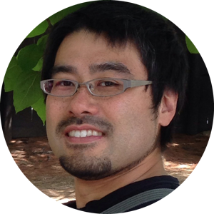 A selfie of Tadatoshi Sato wearing a broad smile in his glasses. He has a short dark hair and a goatee. The photo is set in front of large green leaves as a background.