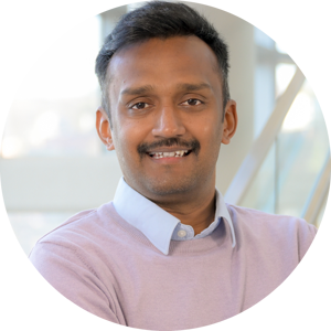 A headshot of Varanasi Siva Karthik, smiling broadly with arms crossed. He is wearing a lavender sweater over a light blue collared shirt, near a staircase with large glass windows in the background.