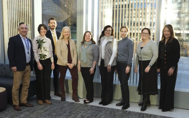 9 individuals standing in front of a large window smiling
